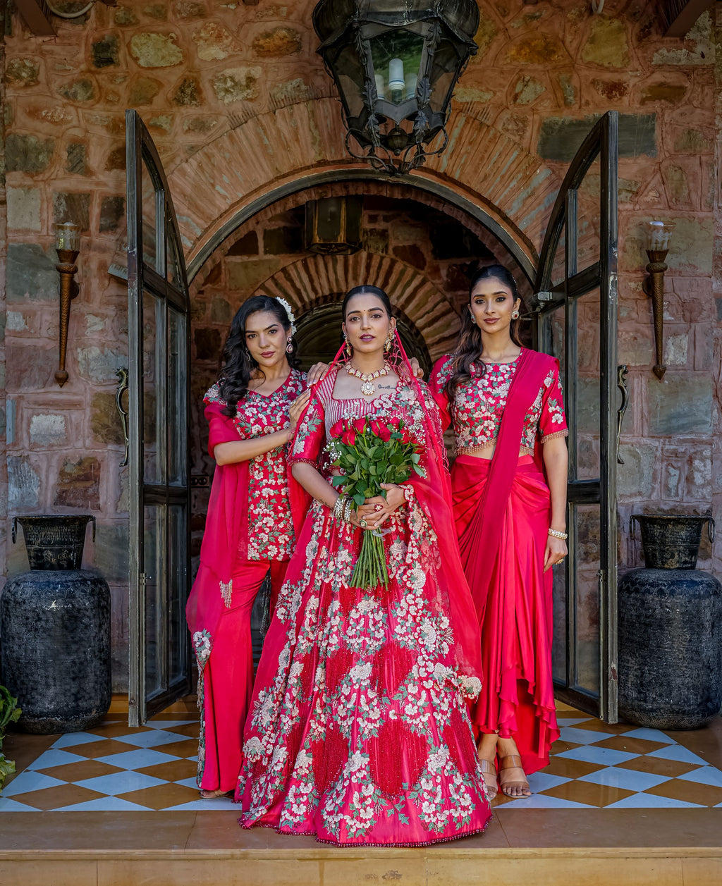 Three women in traditional Indian attire stand in front of an arched stone doorway. The central woman wears a bright pink heavily embroidered lehenga with a matching dupatta and holds a bouquet of red roses, while the two women beside her wear matching pink and red embroidered sari-style outfits.