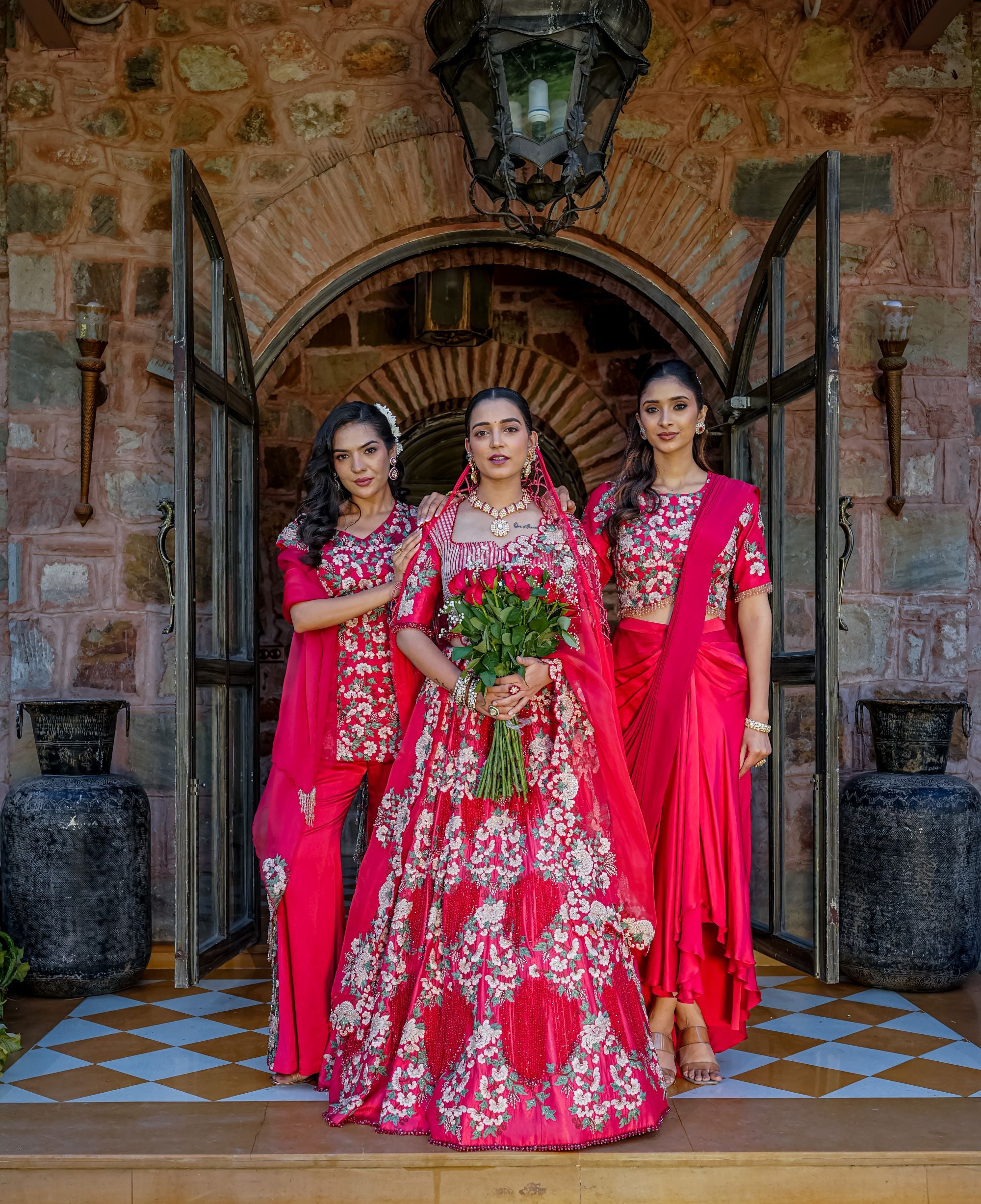 Three women in traditional Indian attire stand in front of an arched stone doorway. The central woman wears a bright pink heavily embroidered lehenga with a matching dupatta and holds a bouquet of red roses, while the two women beside her wear matching pink and red embroidered sari-style outfits.