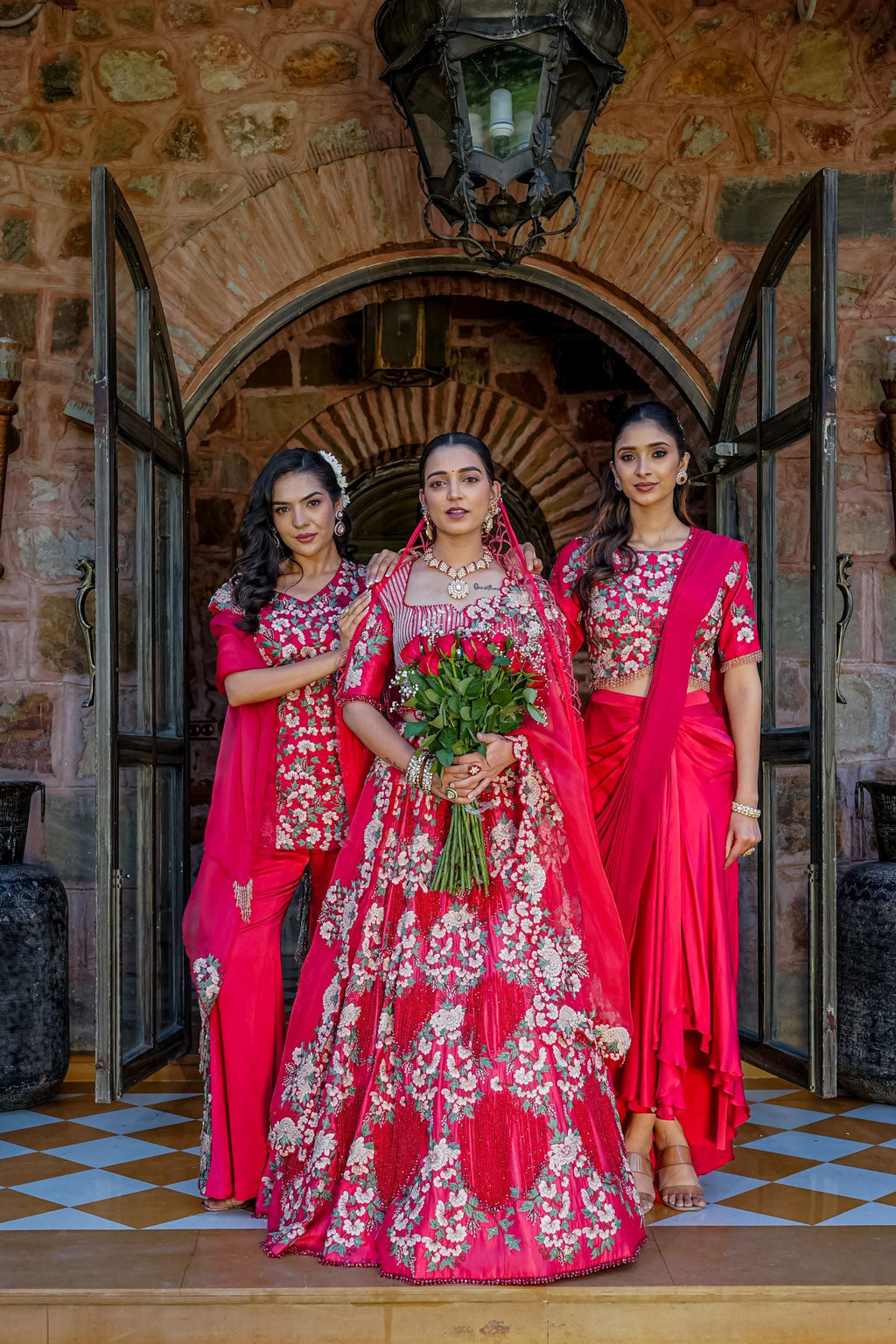 Three women in traditional Indian attire stand in front of an arched stone doorway. The central woman wears a bright pink heavily embroidered lehenga with a matching dupatta and holds a bouquet of red roses, while the two women beside her wear matching pink and red embroidered sari-style outfits.