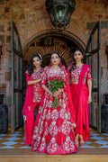 Three women in traditional Indian attire stand in front of an arched stone doorway. The central woman wears a bright pink heavily embroidered lehenga with a matching dupatta and holds a bouquet of red roses, while the two women beside her wear matching pink and red embroidered sari-style outfits.