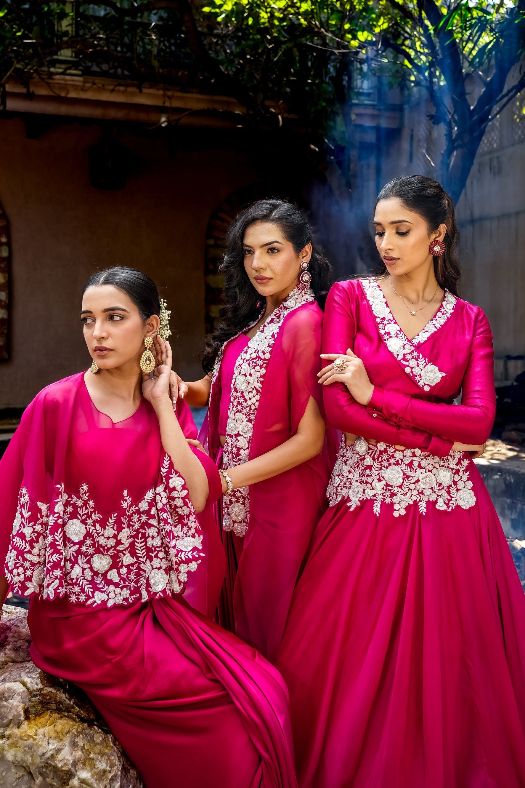 Three women wearing matching bright pink traditional outfits with white floral embroidery, posed outdoors in front of a rustic building with trees in the background.
