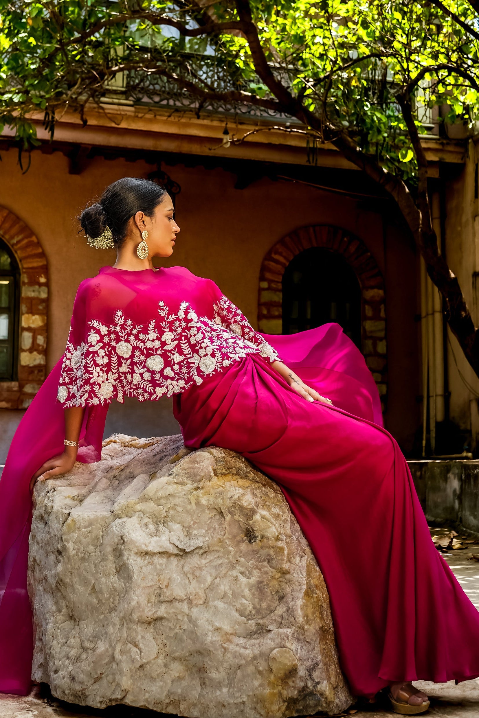 A woman wearing a bright magenta pink traditional outfit with white floral embroidery sits on a large stone in front of a building with arches and leafy trees.