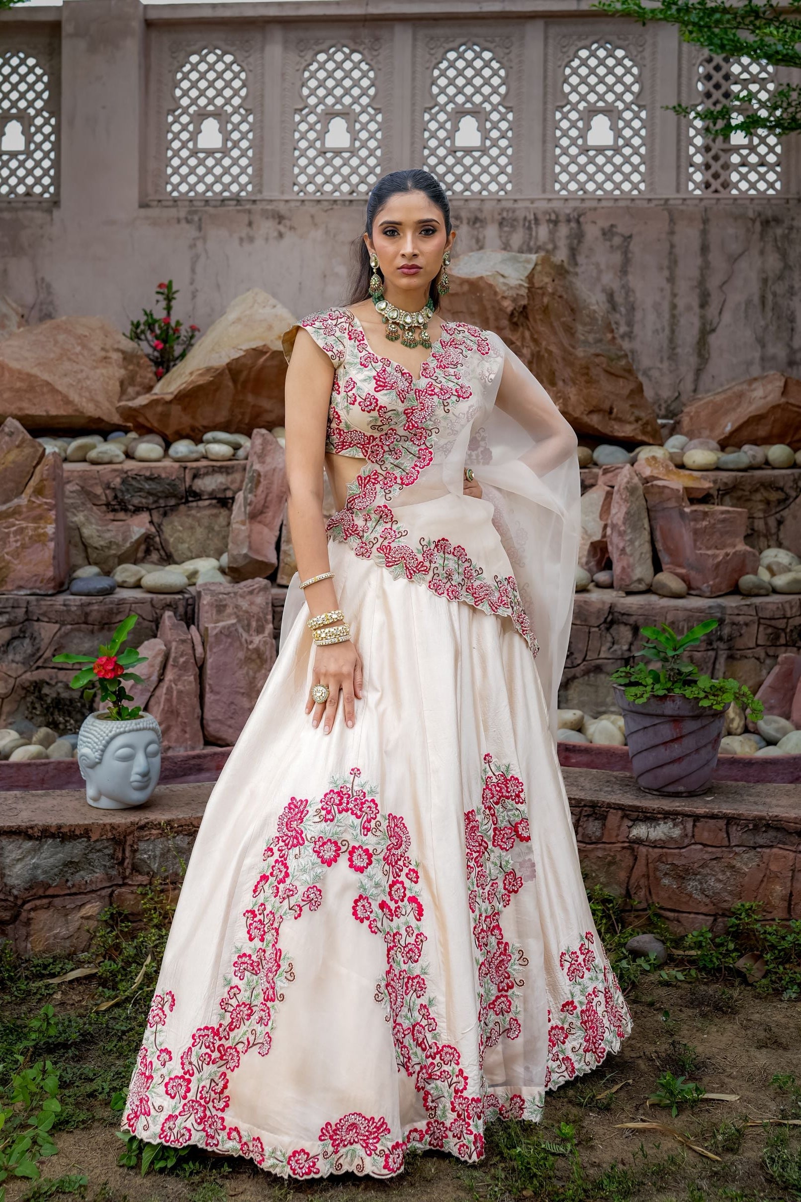 Woman in a white and pink embroidered dress standing in front of a stone wall with decorative elements.