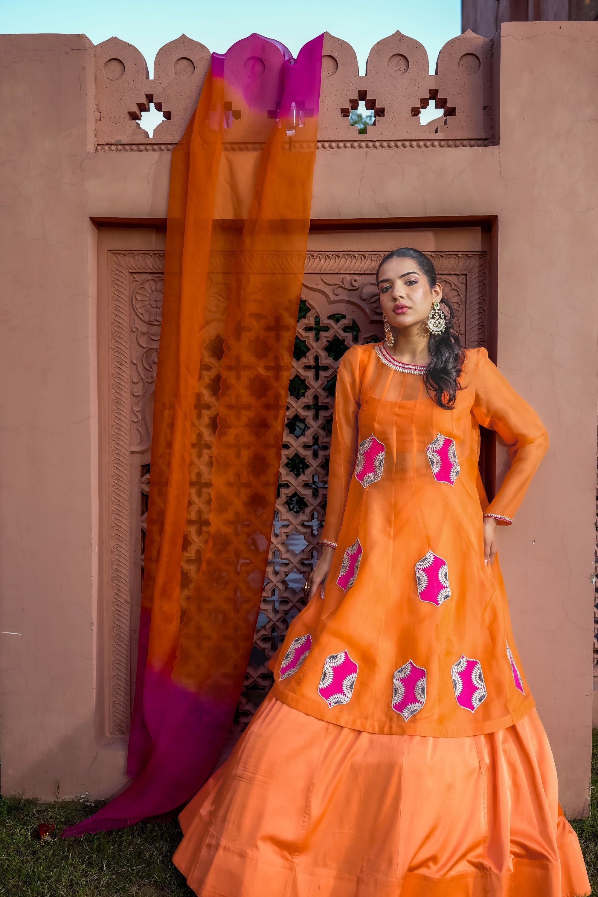 A woman models an orange traditional kurta set with pink and white embroidered motifs, standing in front of a terracotta-colored carved wall with a draped orange dupatta.